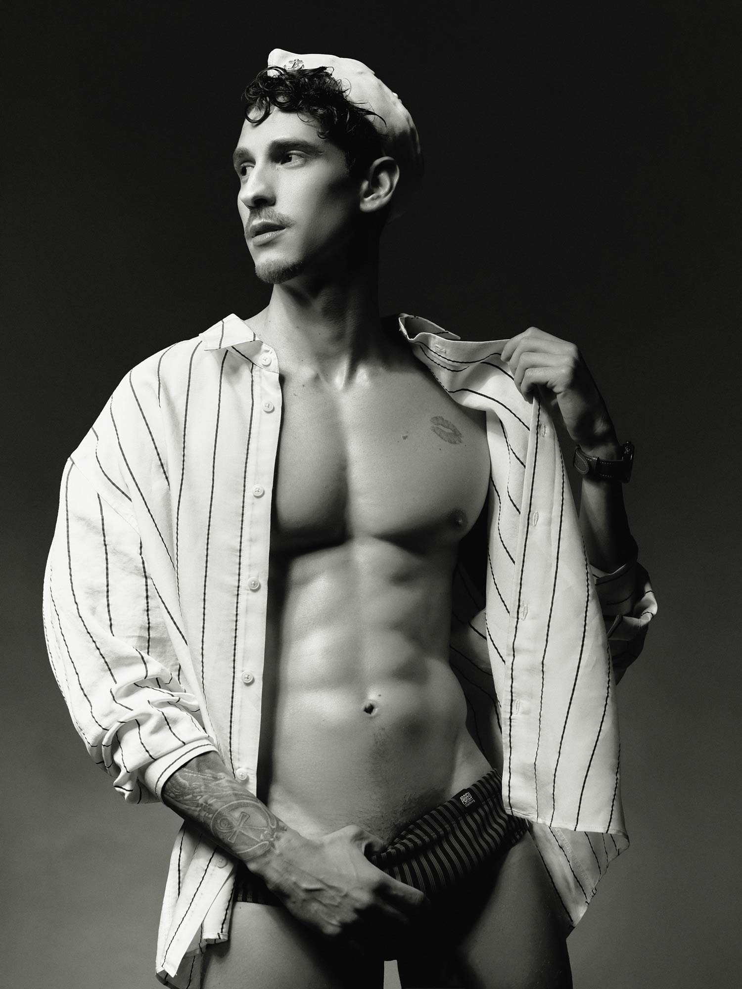A shirtless man in striped pants and cap poses in black and white, partially covering his lower body where the Gabriel Coimbra Dildo by TOYS4FANS—Coming soon—is featured.