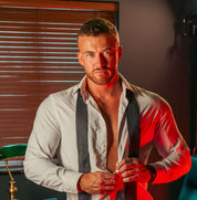 A man with a trimmed beard and short hair, in an unbuttoned white shirt and loosened tie, stands indoors beside the TOYS4FANS Johnny Bravo (Coming soon) silicone dildo, subtly placed on the table under red and natural lighting.