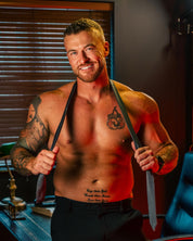 A shirtless, tattooed man poses in a warmly lit office with dark wood blinds, smiling at the camera while showcasing the upcoming TOYS4FANS product, "Johnny Bravo," and wearing a loosened tie around his neck.