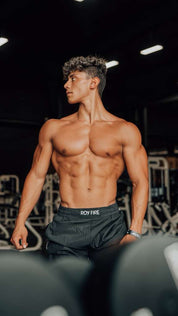 A muscular young man with defined abs and curly hair stands shirtless in a gym, wearing black shorts. Behind him is exercise equipment. Josue Fit by TOYS4FANS—coming soon!.