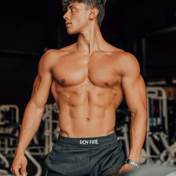 A muscular young man with defined abs and curly hair stands shirtless in a gym, wearing black shorts. Behind him is exercise equipment. Josue Fit by TOYS4FANS—coming soon!.