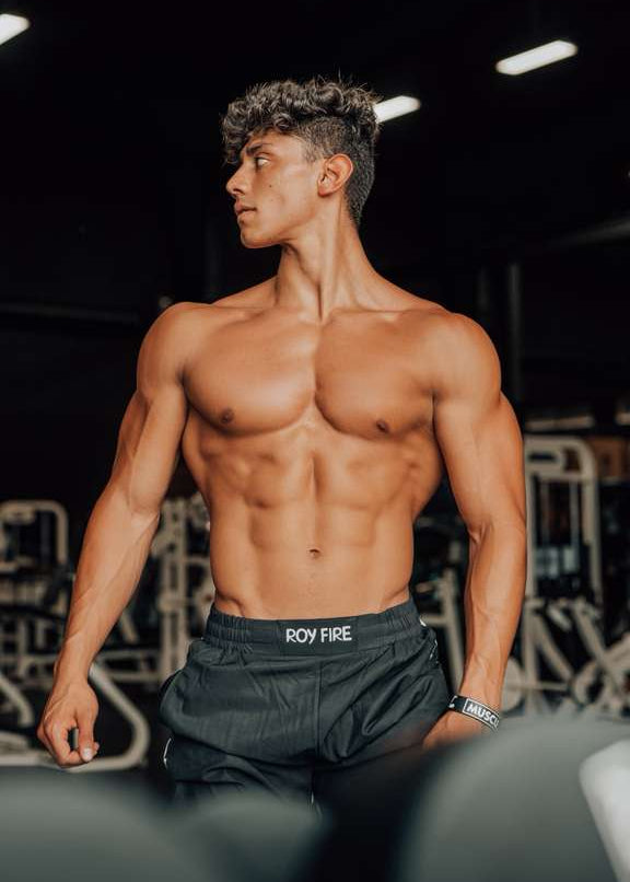 A muscular young man with defined abs and curly hair stands shirtless in a gym, wearing black shorts. Behind him is exercise equipment. Josue Fit by TOYS4FANS—coming soon!.