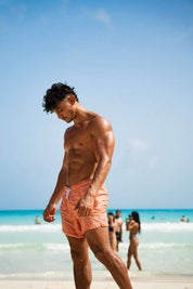 A shirtless man in pink swim trunks by TOYS4FANS stands on a sunny beach, gazing down as the blue ocean and sky create a vibrant scene—perfect for highlighting the lifelike Josue Fit figure, coming soon.